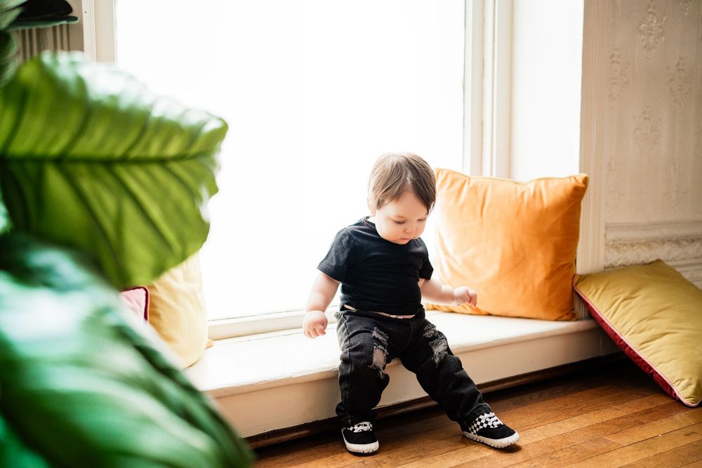 Curious toddler in black outfit stands at window seat with colorful cushions in bright room.