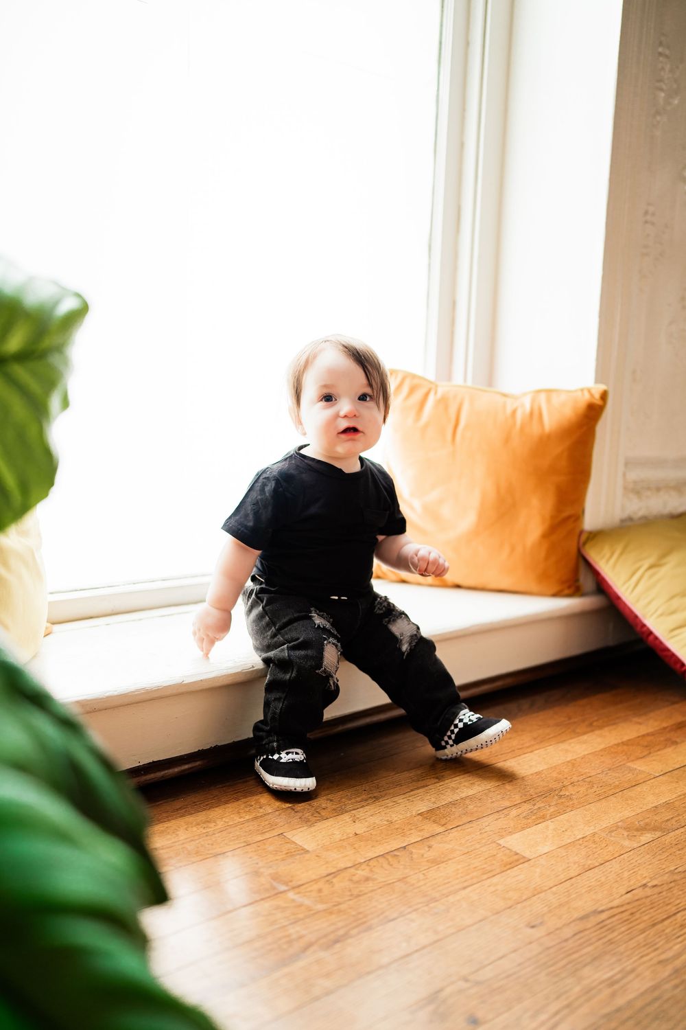 A curious baby in black outfit and sneakers stands on hardwood floor near a bright window with orange cushions.