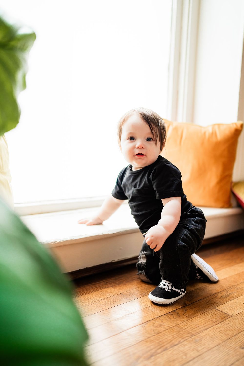 Adorable baby in black clothes and sneakers crouches near a sunlit window seat with colorful cushions indoors.