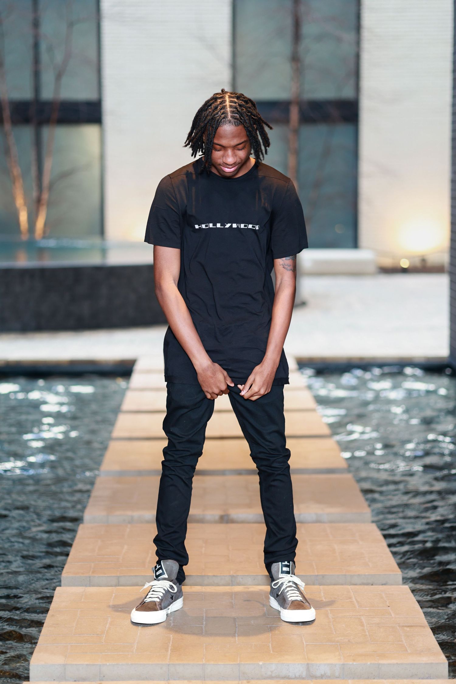 Young man with dreadlocks wearing all-black outfit standing on stepping stones over water feature.