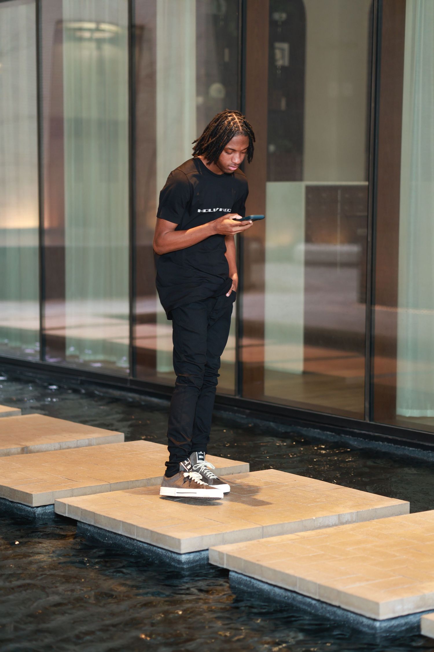 Young Black man with dreadlocks in black outfit uses smartphone while standing on stepping stones over water.