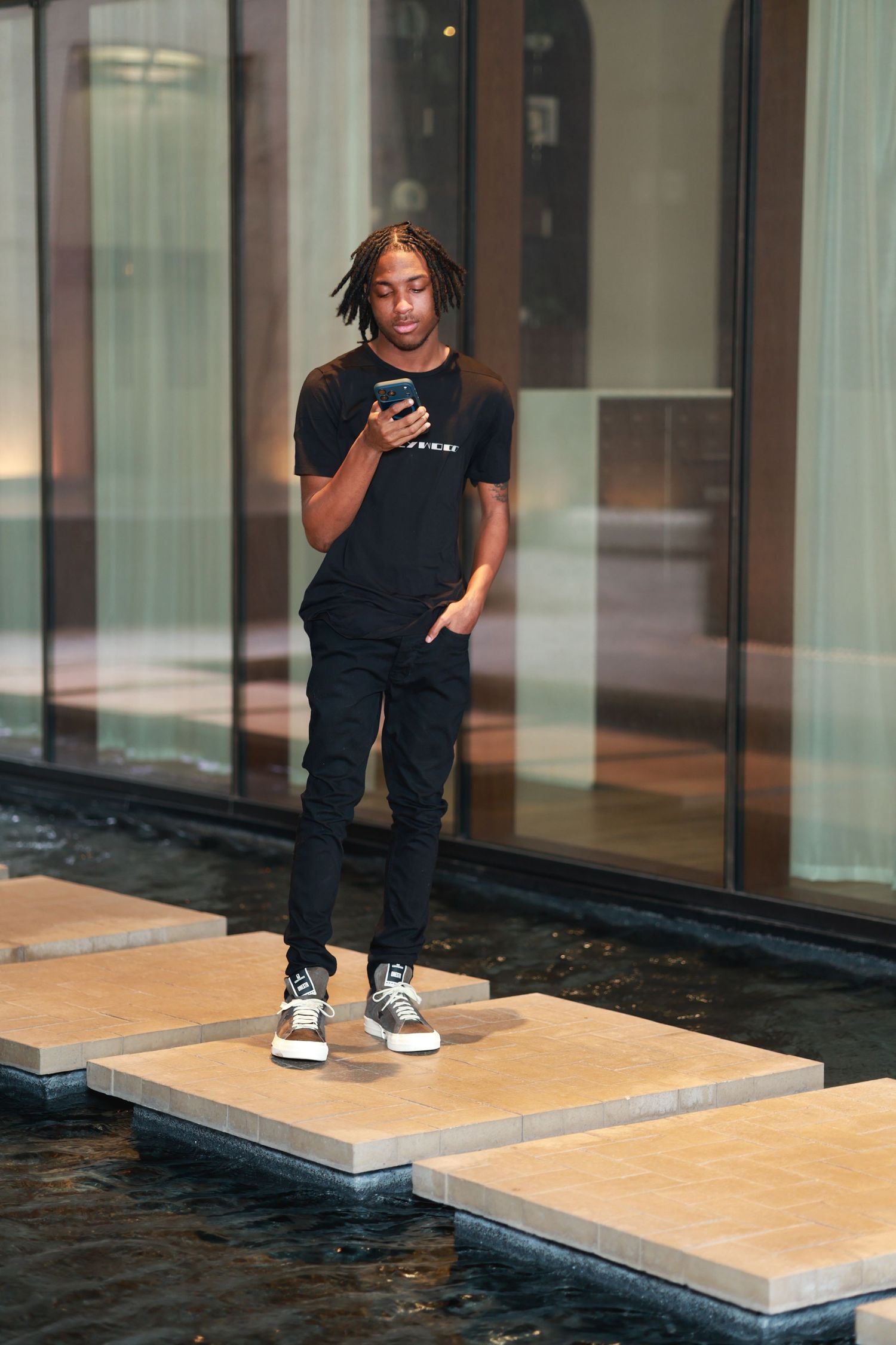Young Black man in all-black outfit standing on stepping stones, looking at smartphone outside modern building.