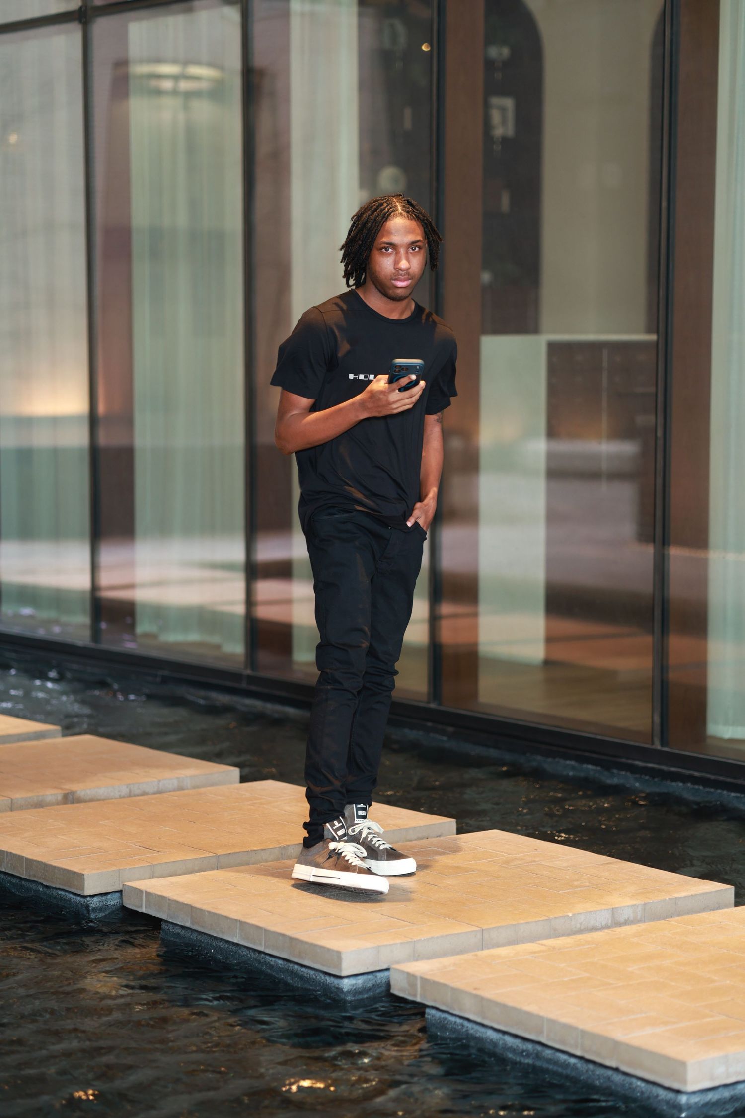 Young Black man in all-black outfit standing on stepping stones over water, holding smartphone outside modern building.