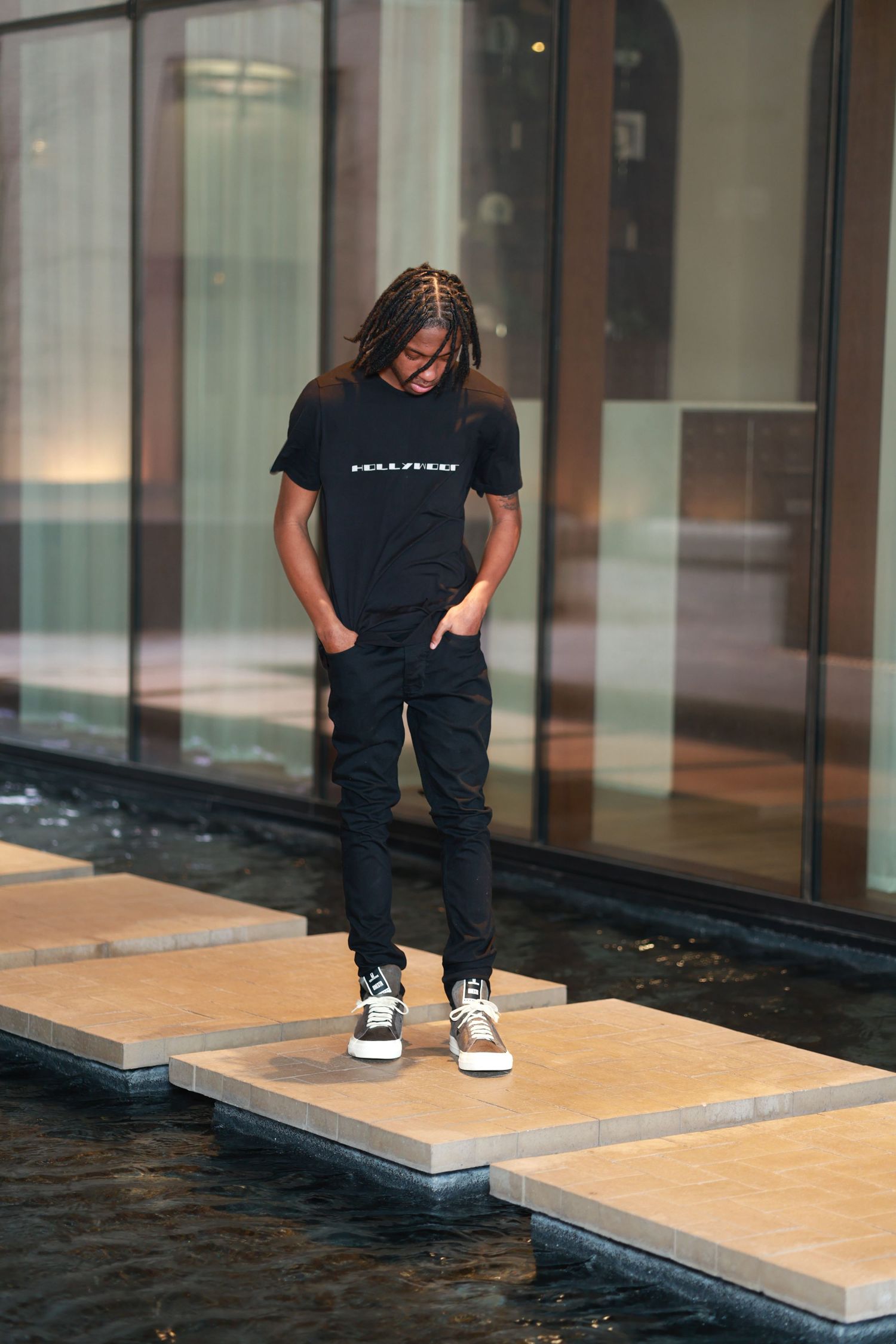 Young man with braids in black t-shirt and jeans standing on stepping stones over water feature.