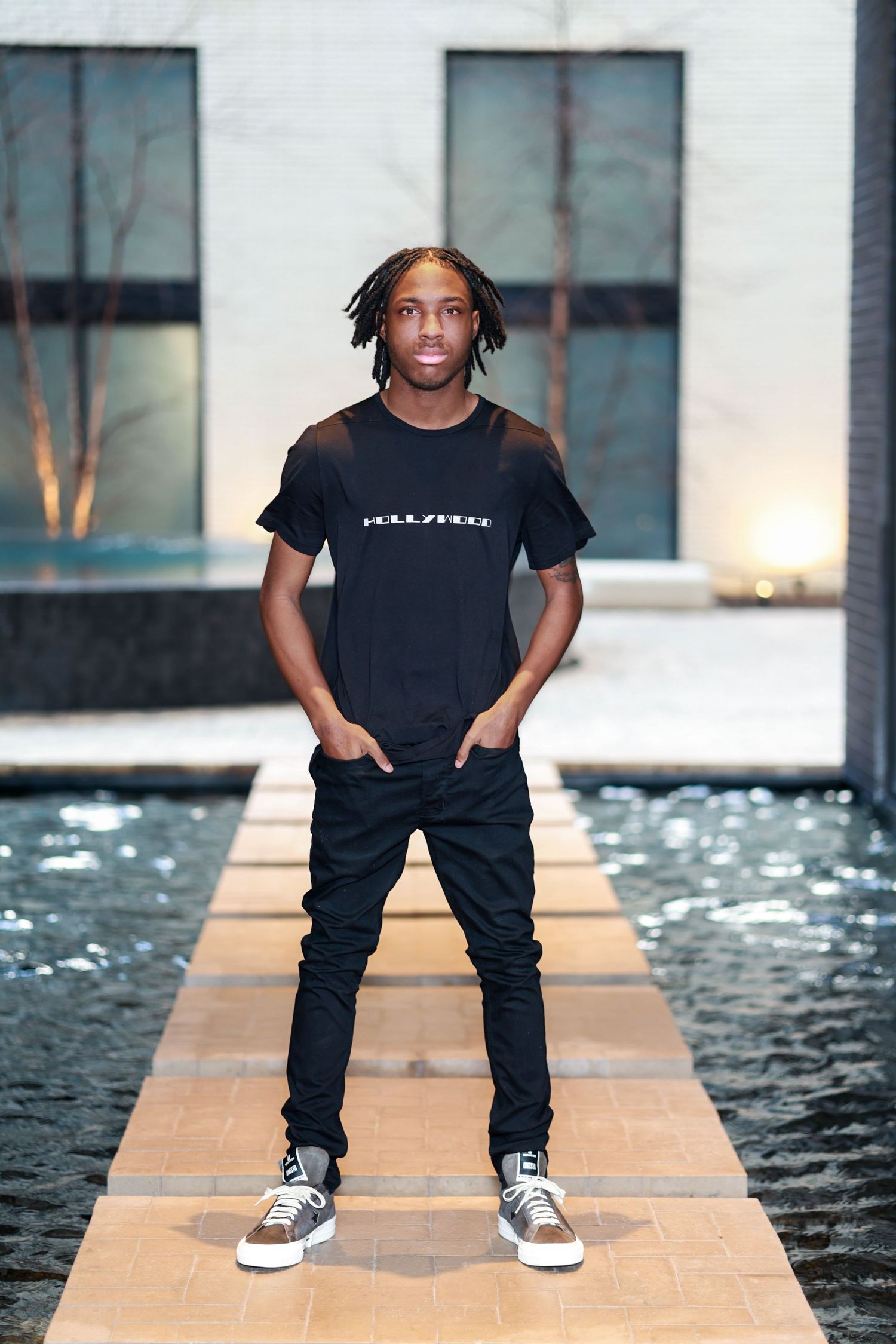 Young man in black Hollywood tee and jeans stands confidently on stone path beside reflective pool at dusk.
