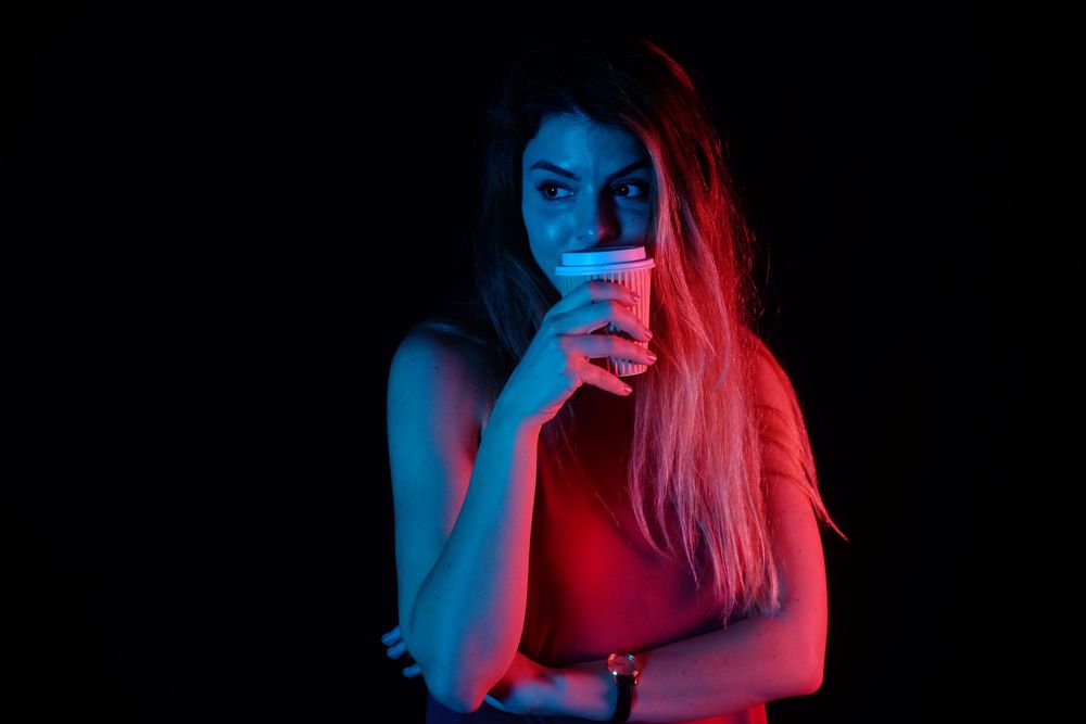 Young woman holding a cup under dramatic red and blue neon lighting against a dark background.