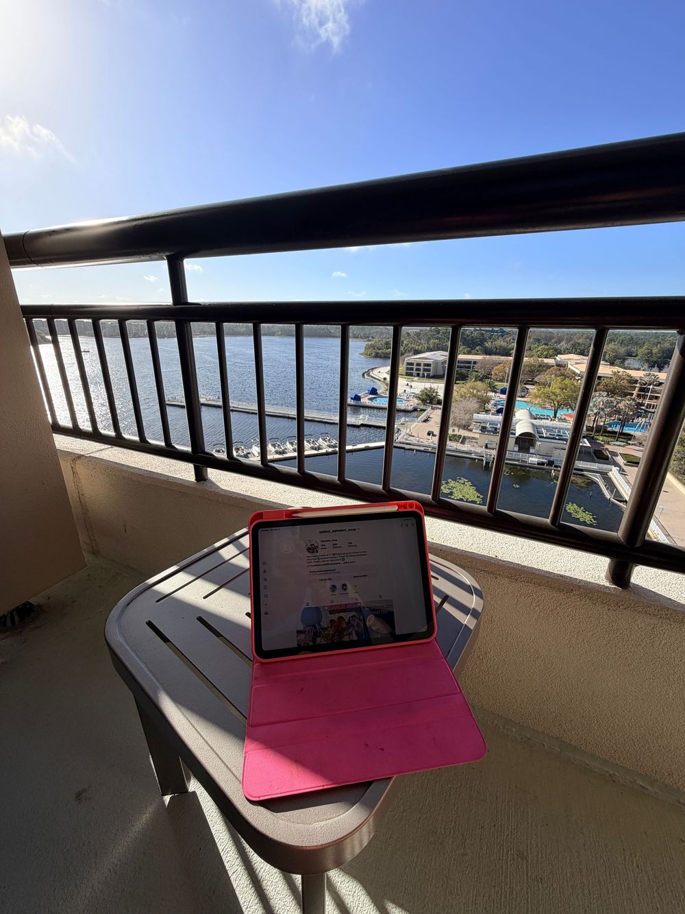 Tablet with pink case sits on balcony table overlooking marina and waterfront view on sunny day.