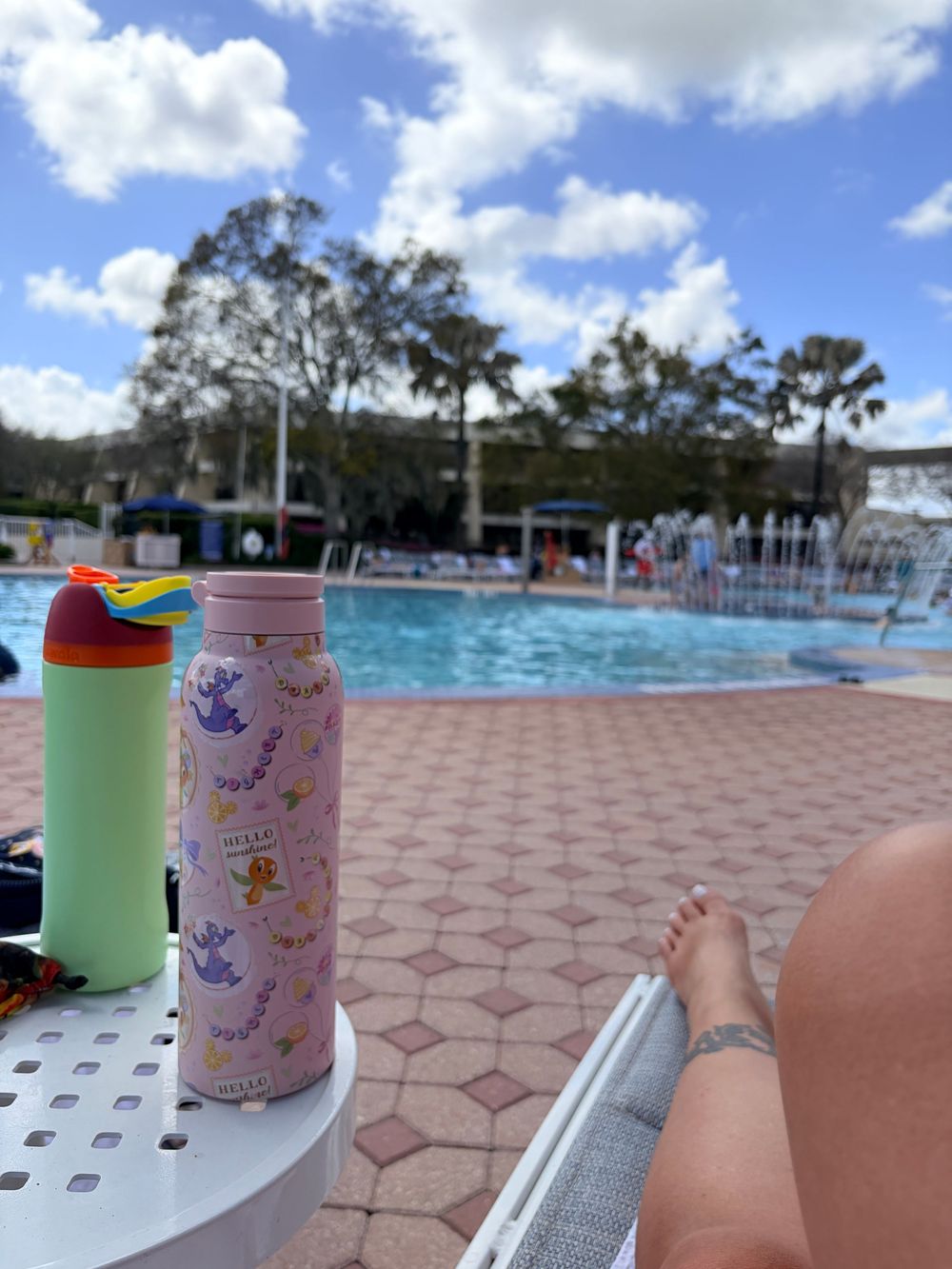Poolside relaxation scene with water bottles and feet visible on a lounge chair near a swimming pool on a sunny day.