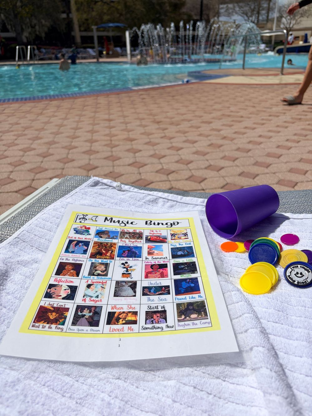 Memory game cards and colorful tokens arranged on a white towel by a swimming pool on a sunny day.