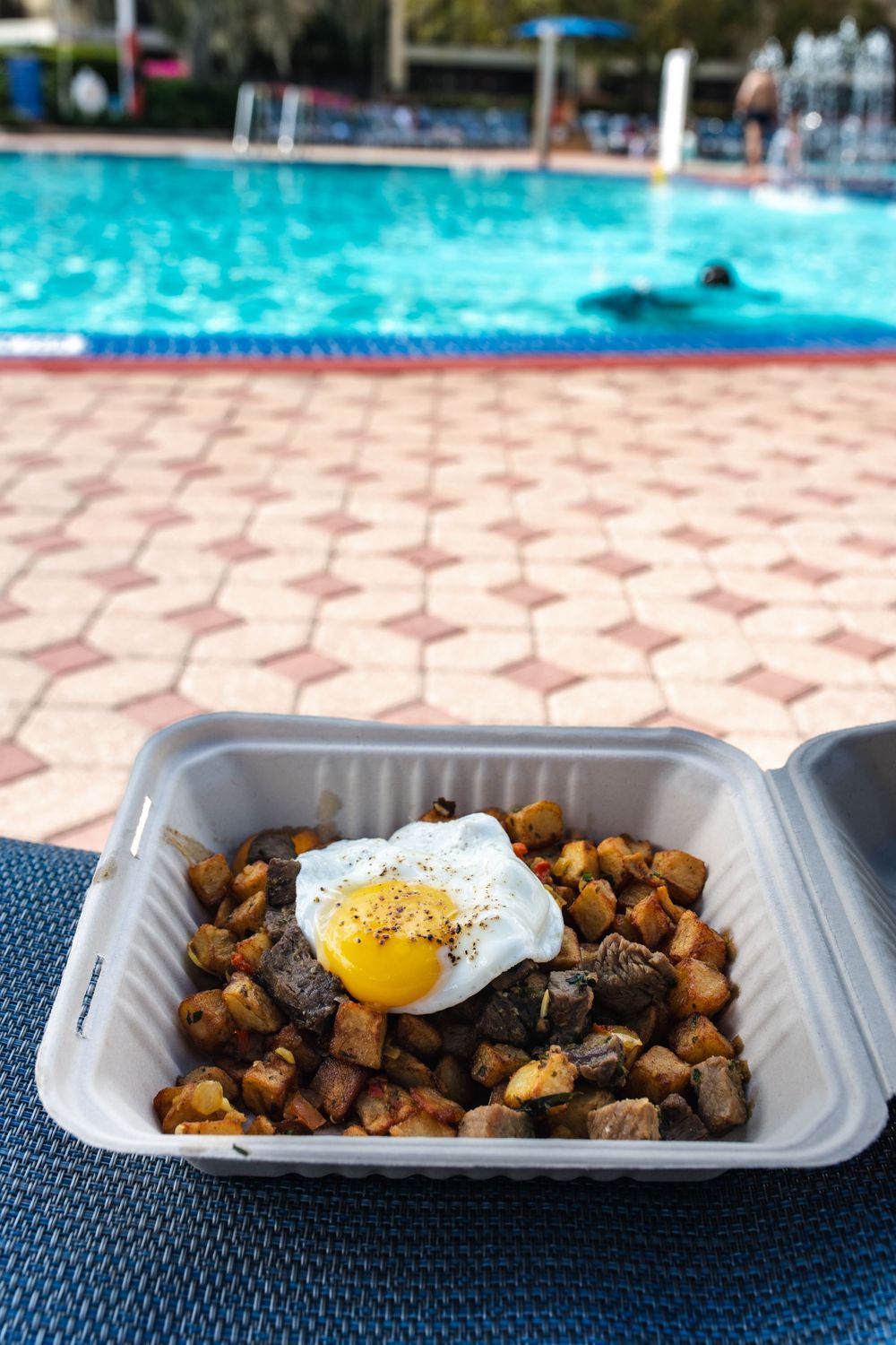 Sunny side up egg served over crispy seasoned breakfast potatoes on a white disposable plate by a pool.