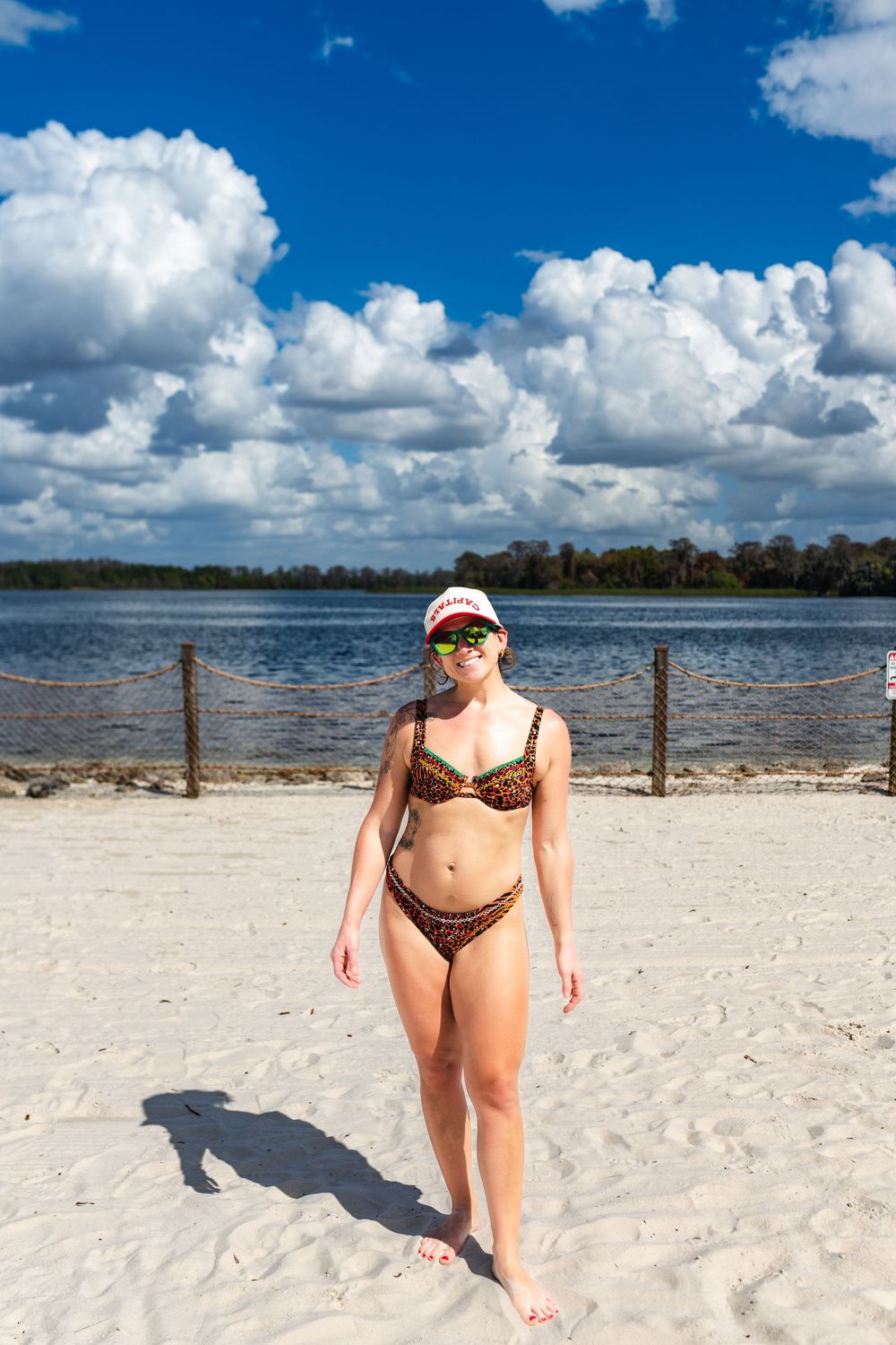 A person in a leopard print swimsuit walks along a pristine white sand beach with blue water and sky in the background.