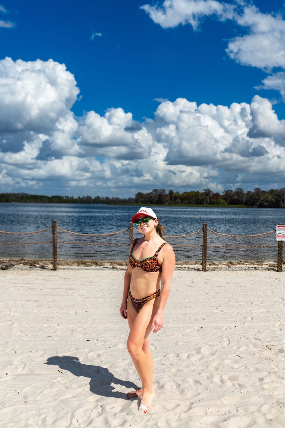 A person in a red cap walks along a sandy beach with blue water and white clouds in the background.