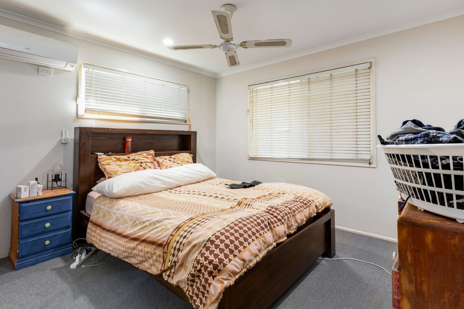 Carpeted bedroom with dark timber bed frame, ceiling fan, split system air conditioner, and venetian blinds.