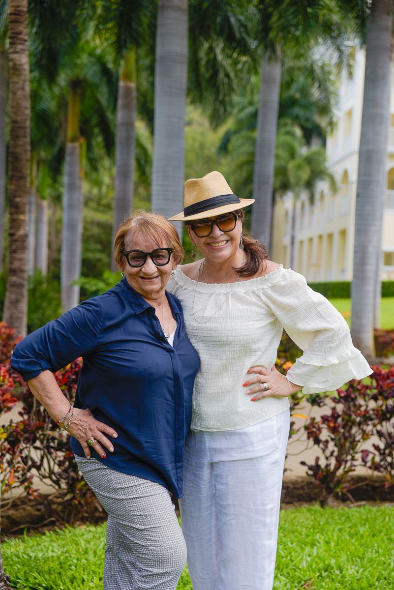 Two people wearing blue and white outfits embrace while standing on grass with palm trees in the background.