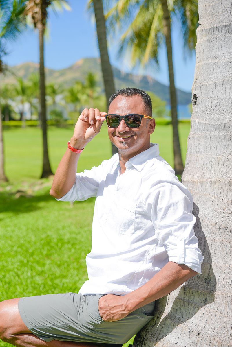 Person in white shirt and sunglasses relaxes against palm tree on sunny tropical lawn with mountains in background.
