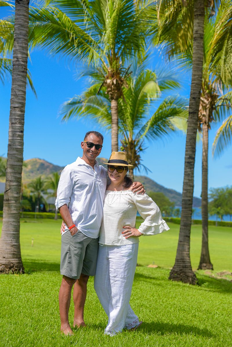 Couple in white outfits walking through a tropical palm tree garden with mountains in the background on a sunny day.