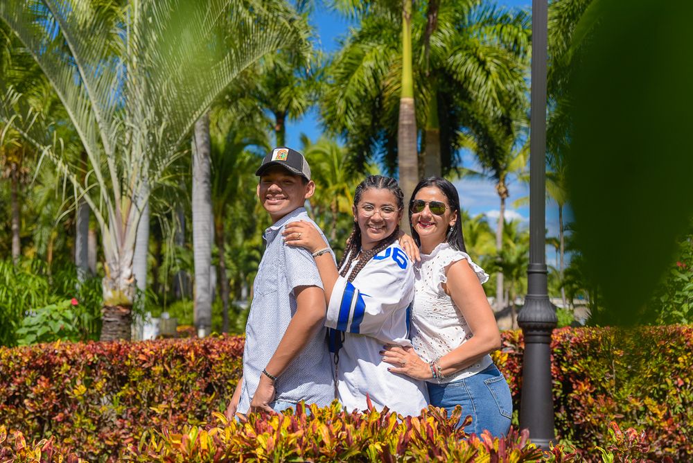 Three people pose together in a tropical garden with palm trees and colorful flowers in the background.