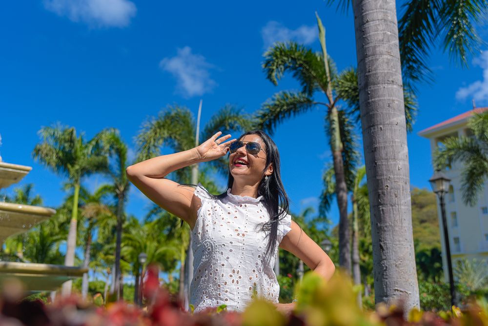 A person in a white top poses joyfully against a backdrop of tall palm trees and bright blue sky.
