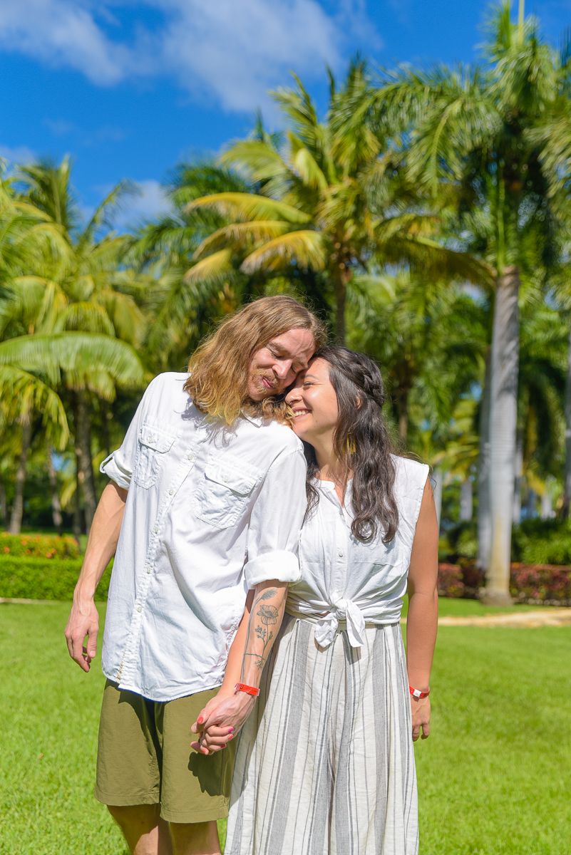 Couple in white outfits sharing a tender moment on a palm tree-lined lawn in a tropical setting.