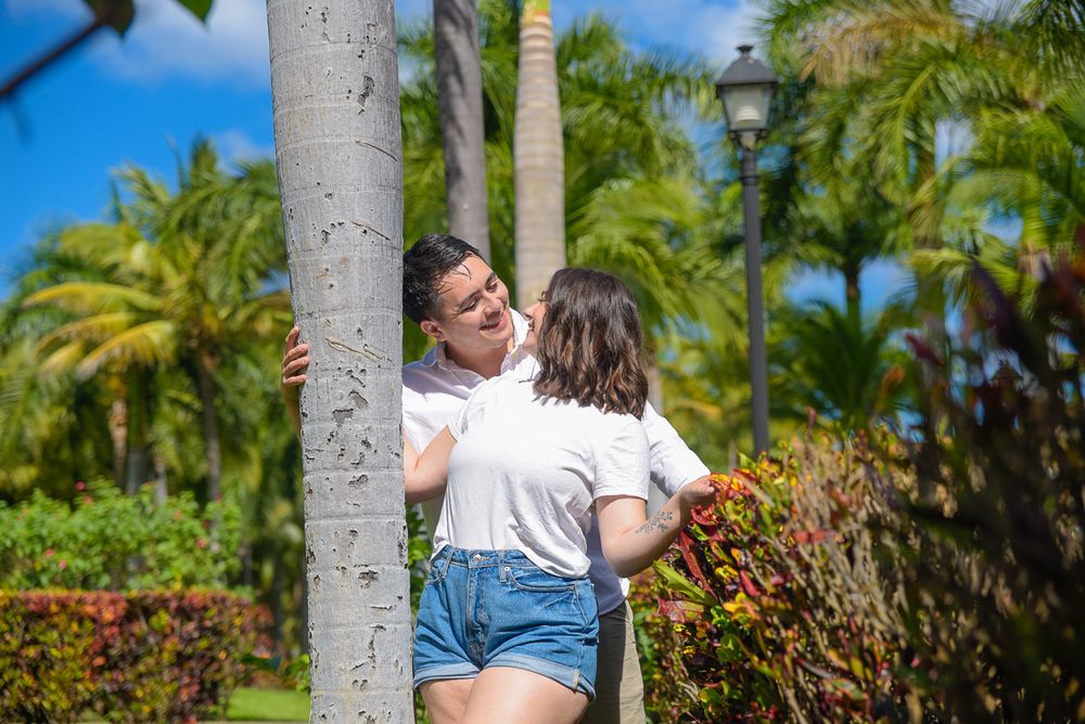 Two people share a playful moment by a palm tree in a lush tropical garden setting.
