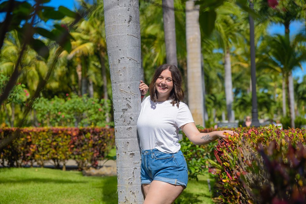 Person in denim shorts and white t-shirt poses near palm trees in a tropical garden setting.
