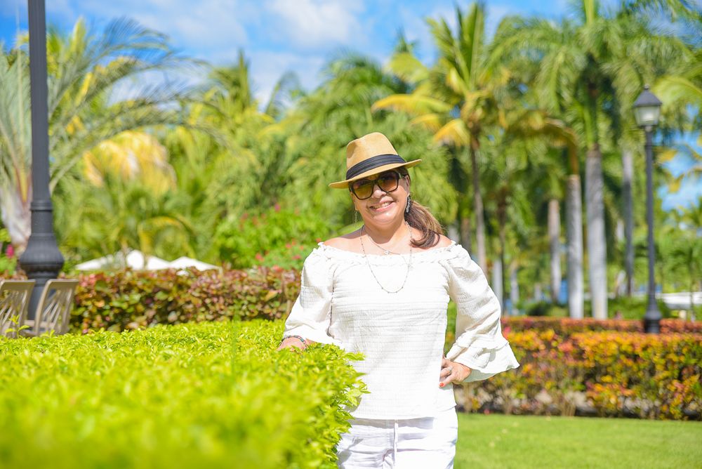 Person in white clothing and sun hat poses in front of tropical garden with palm trees.
