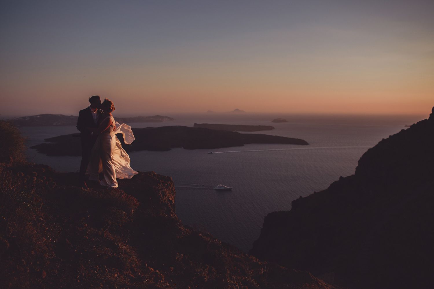 Santorini destination wedding photographer capturing couple on cliffs overlooking the caldera at sunset.