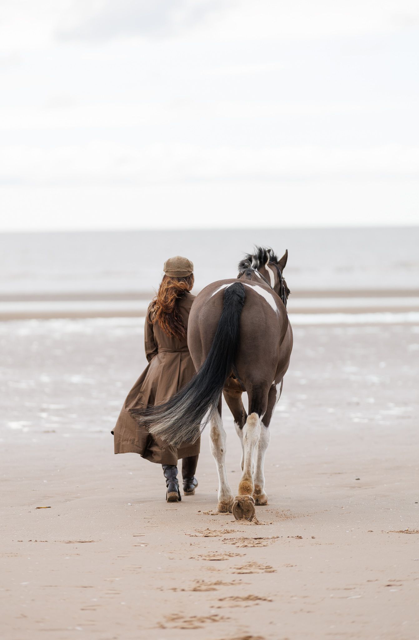 A person walks along a misty beach with a majestic brown and white horse on a cloudy day.