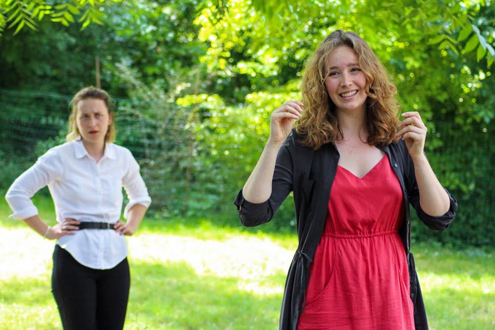Two young women stand outdoors in a green garden, one in red dress and one in white shirt.