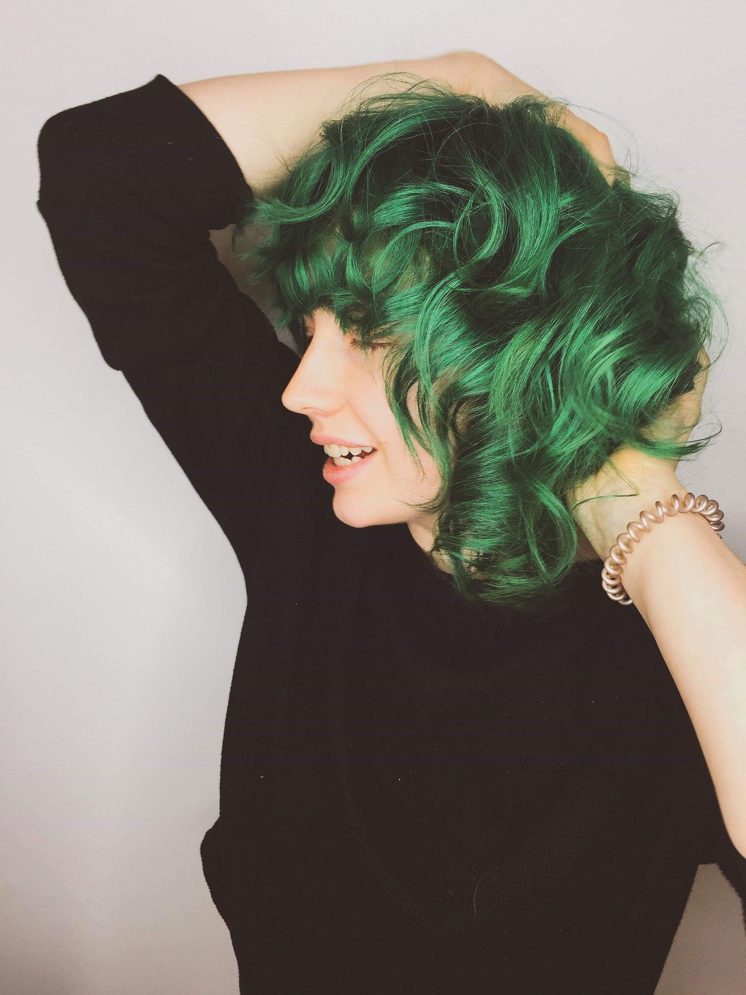 Woman with vibrant green curly hair poses in profile view wearing a black top with bracelet.