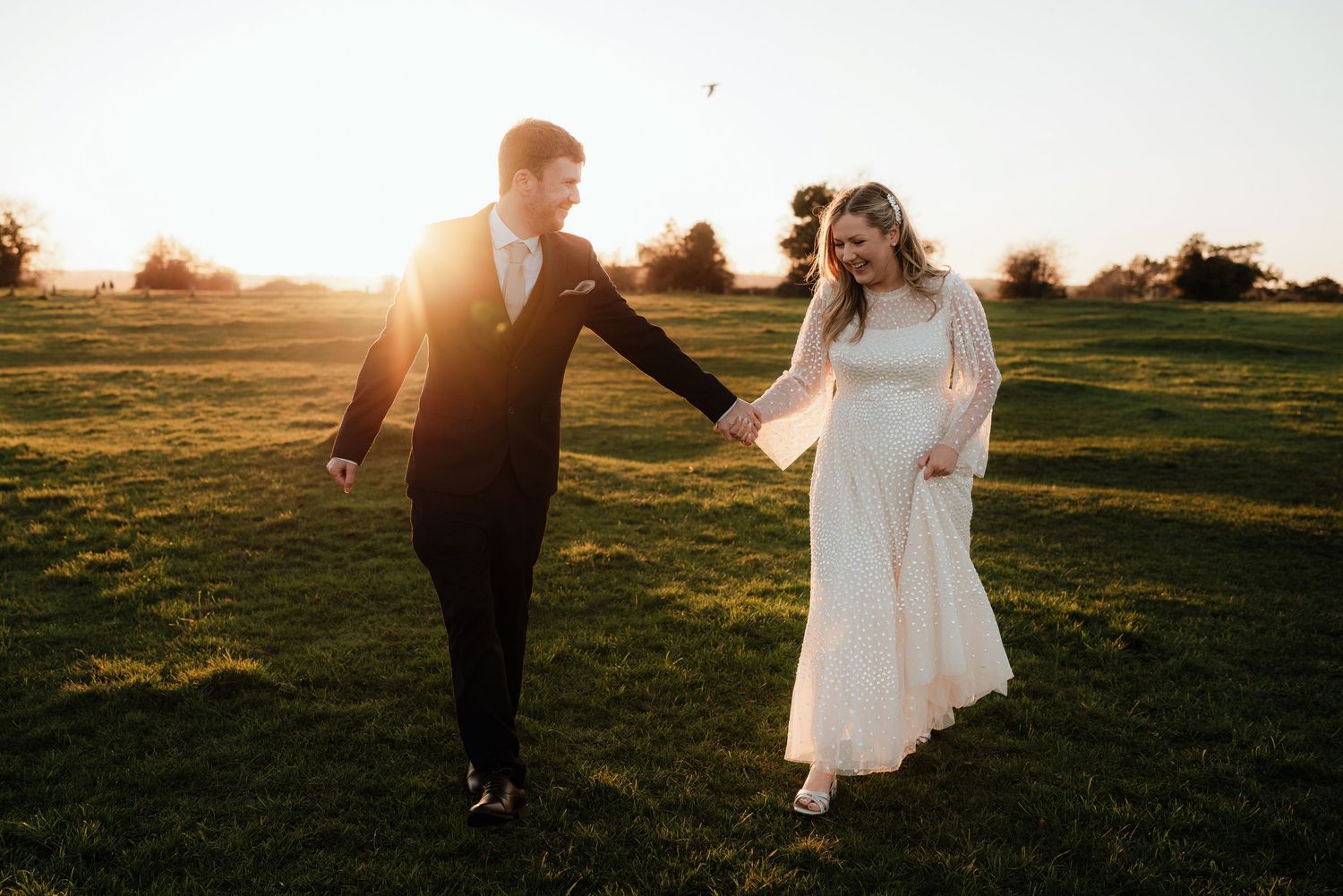 A couple walking together in a field at sunset, the woman wearing a long white dress and the man in a dark suit.