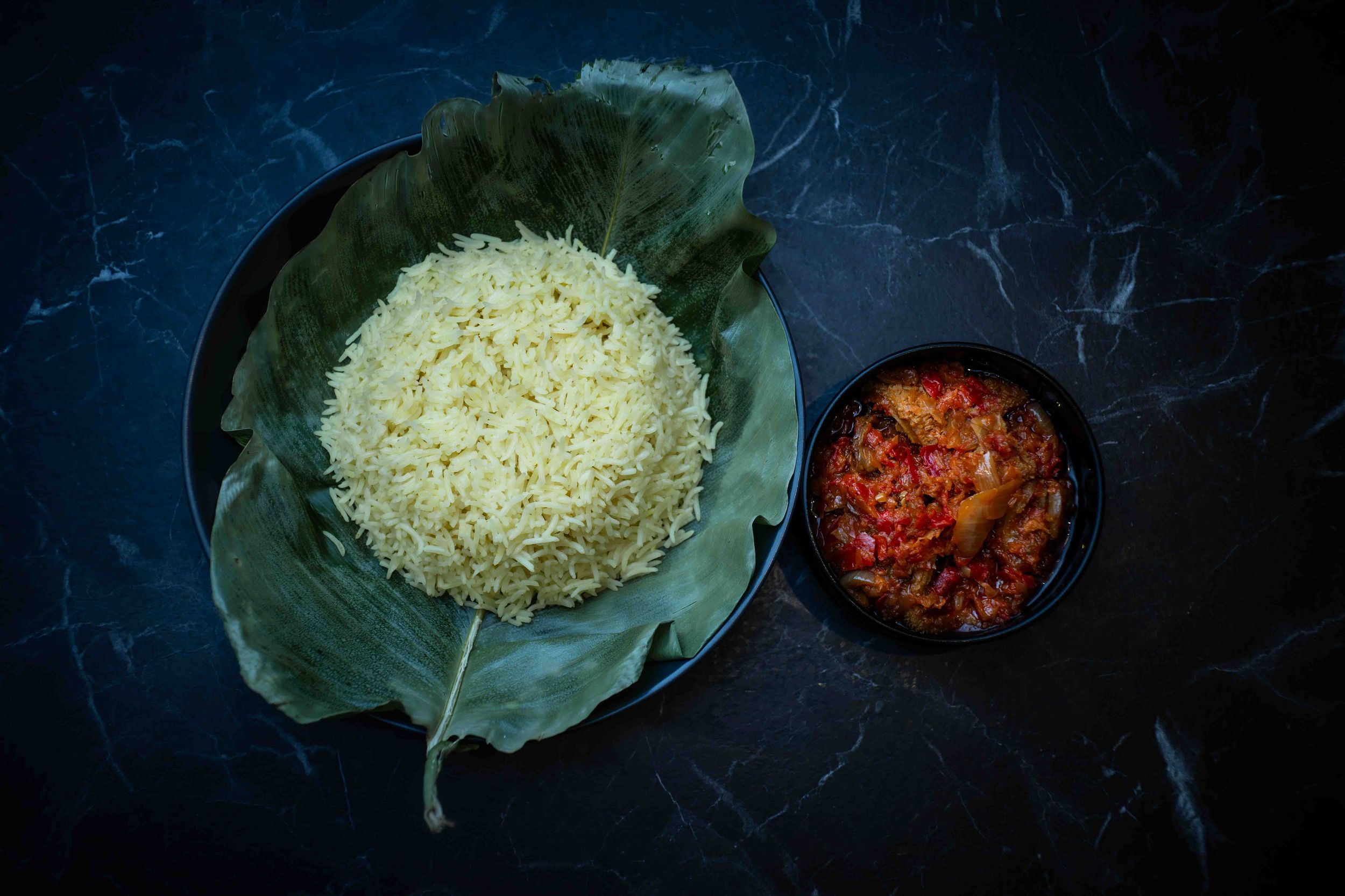 Steamed white rice served on banana leaf with spicy red sambal chili sauce in black bowl on dark marble surface.