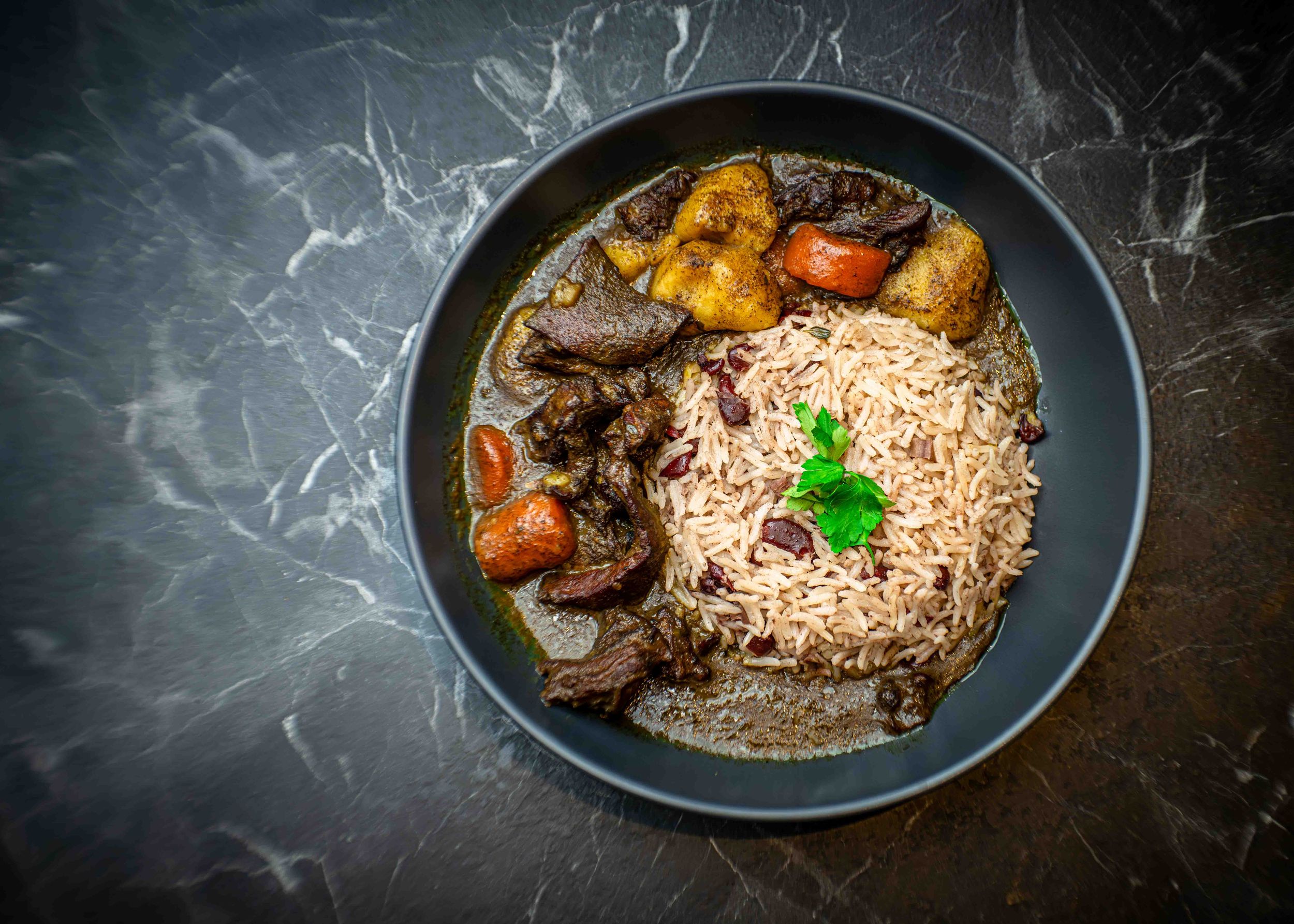 A bowl of rice and peas with braised oxtail, fried plantains, and tomatoes garnished with fresh herbs.