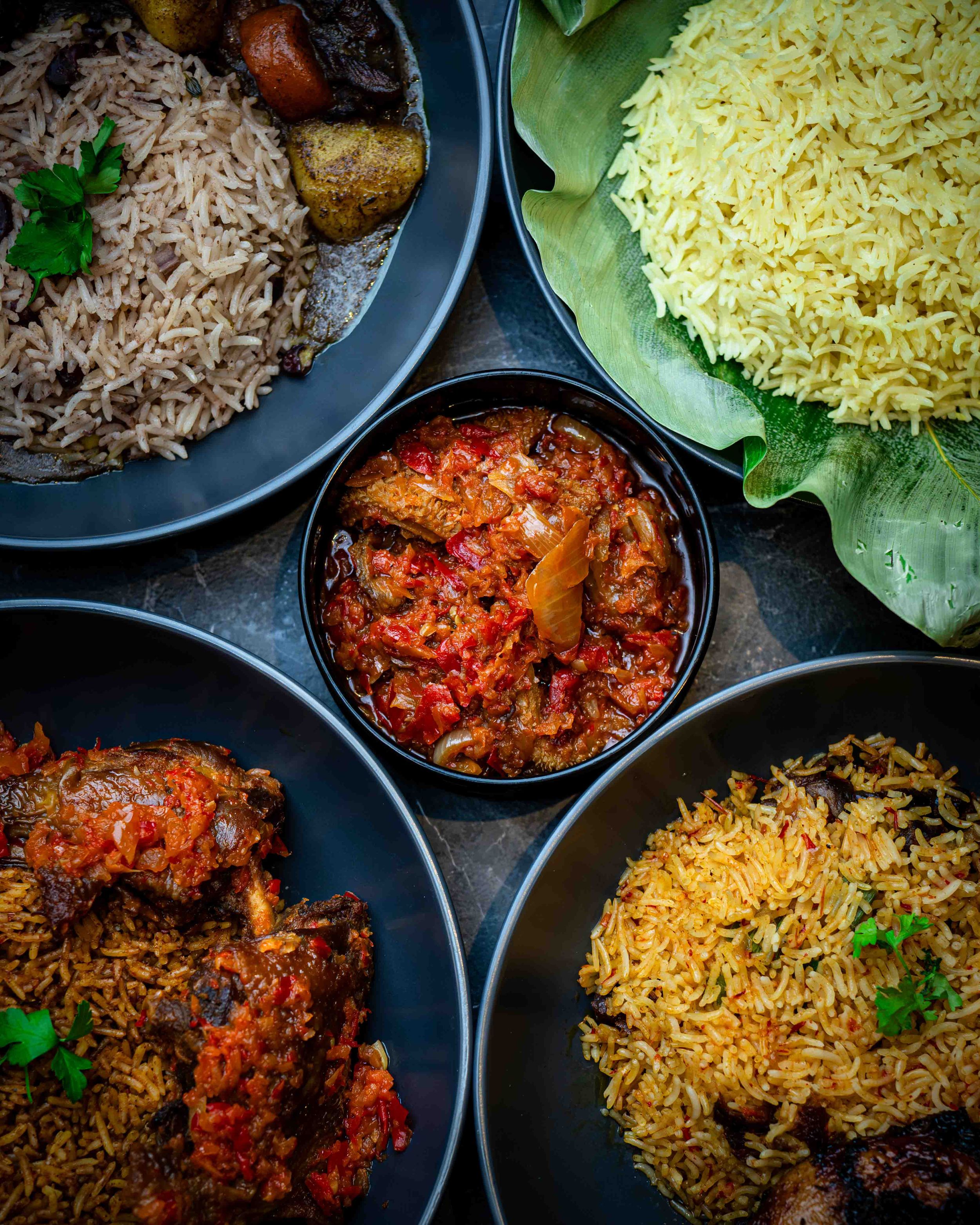 Overhead view of African rice dishes with tomato stew, plantains, and garnished plates on dark background.