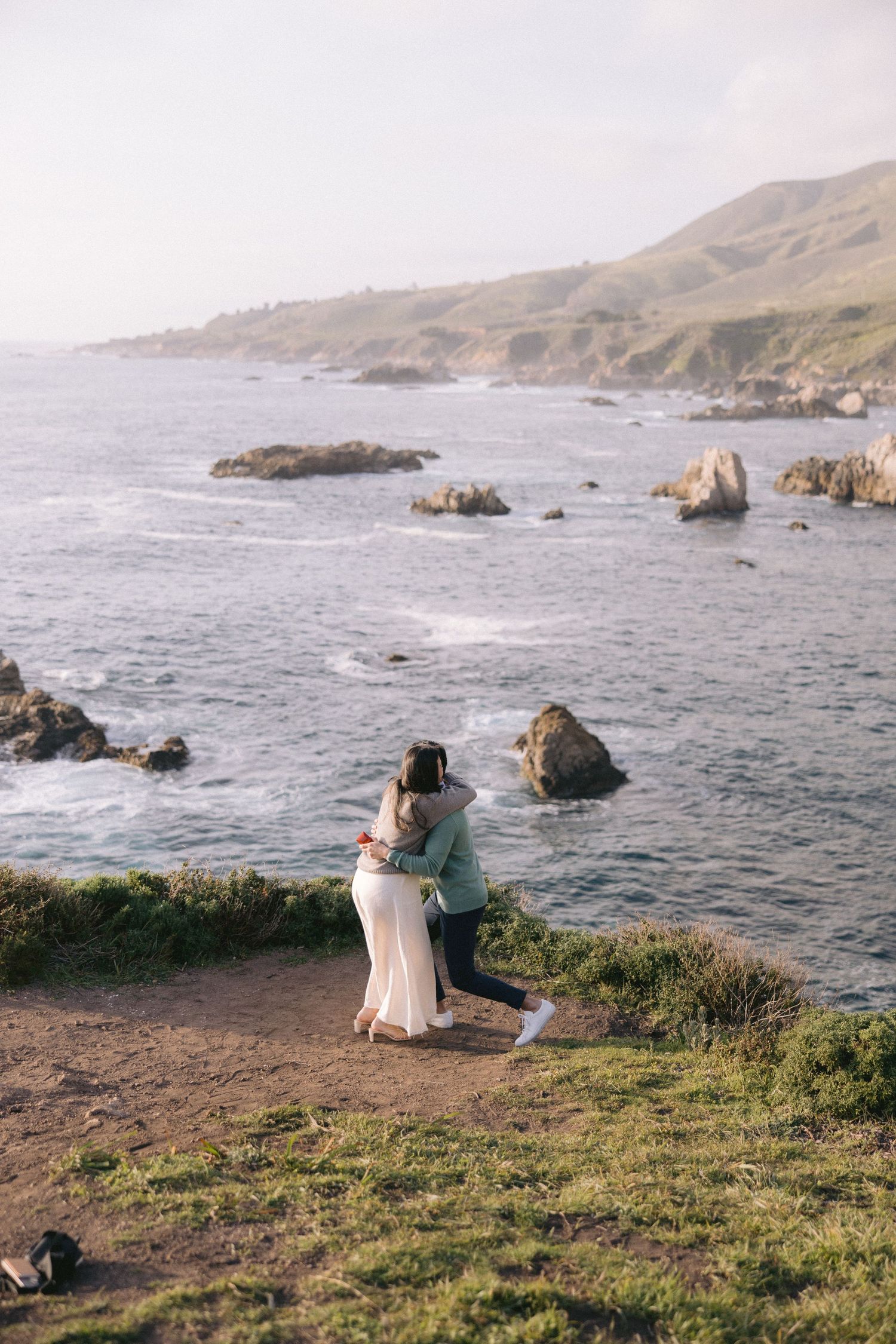 Two figures embrace on a cliff overlooking the dramatic Big Sur coastline with rocky outcrops and waves below at sunset.