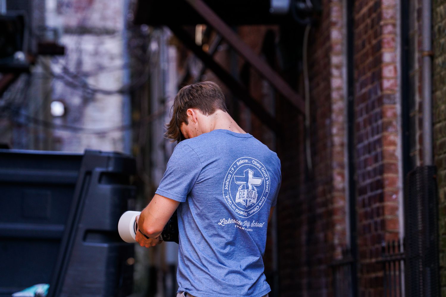 Person wearing blue t-shirt with cross logo stands near brick building.