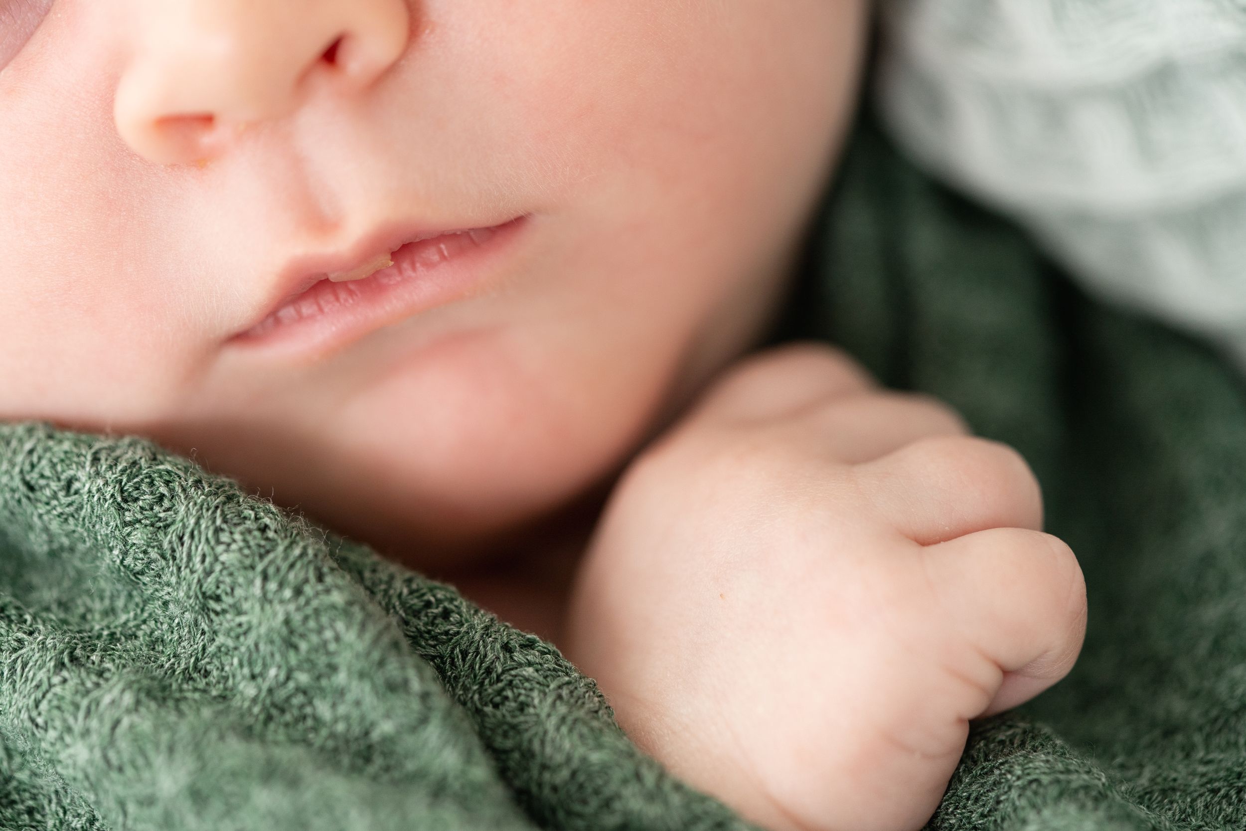 A baby rests peacefully in a soft sage green blanket, showing a tiny hand and a partial view of their face.