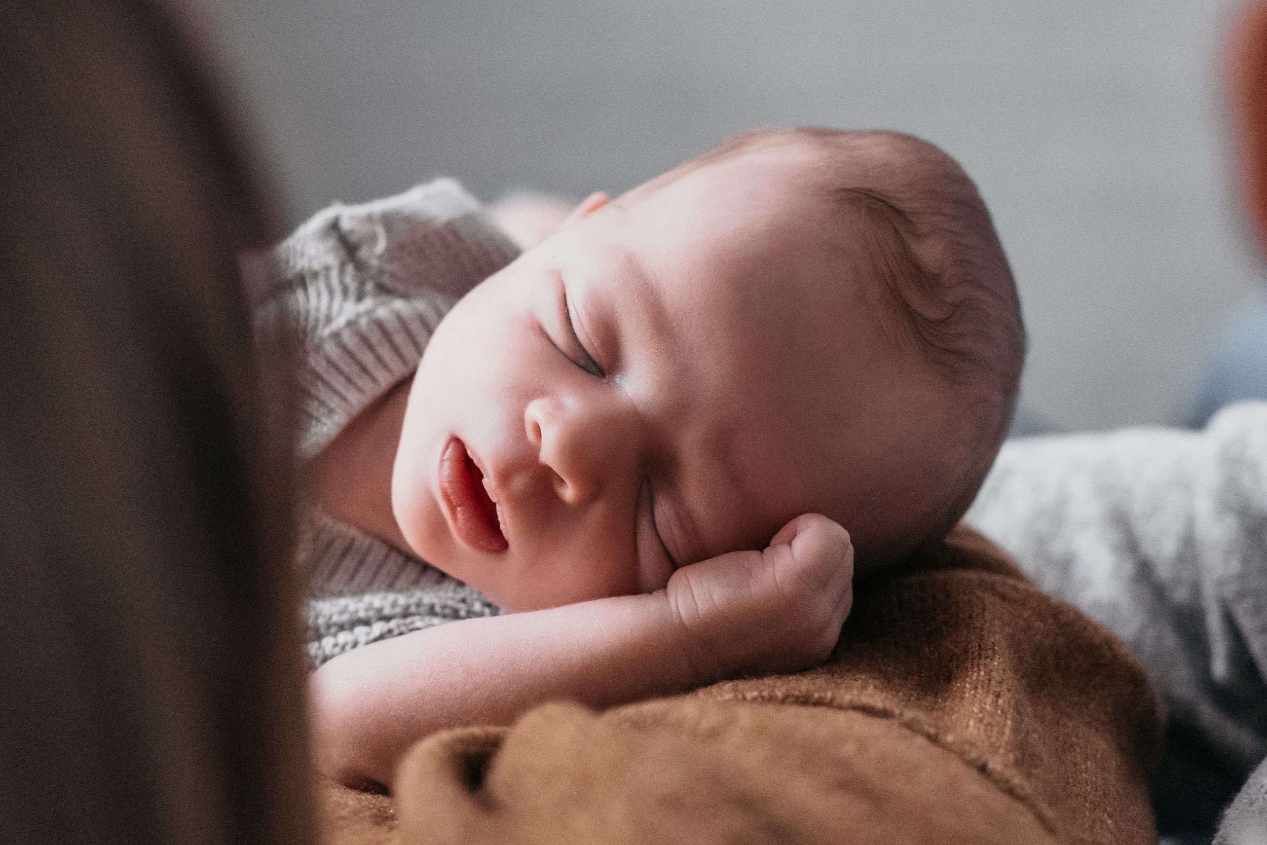 A peaceful sleeping baby rests comfortably on a brown blanket in soft natural lighting.