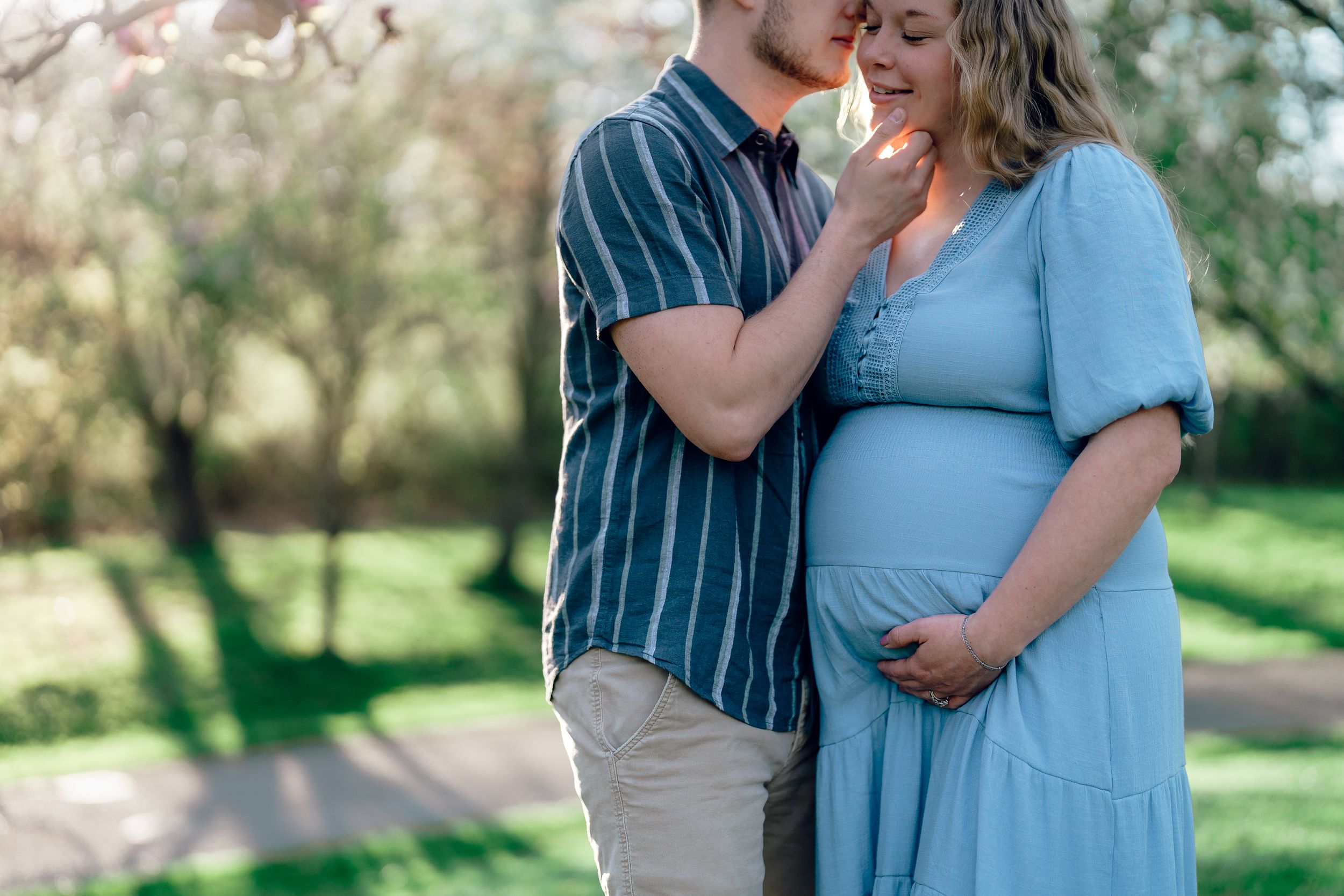 A couple shares an intimate moment in a sunlit garden setting while wearing coordinating blue outfits at maternity photo session in Akron OH