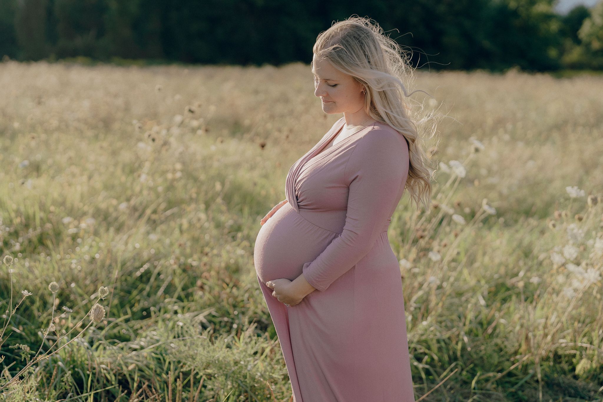An expectant mother stands in profile wearing a pink dress in a sunlit golden meadow field.