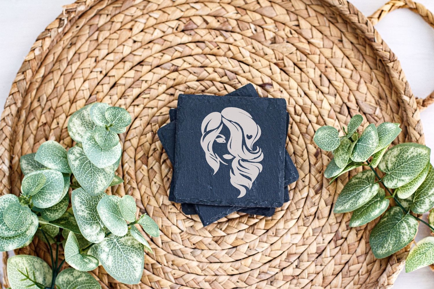 Black decorative card with silver design placed on woven rattan tray with green eucalyptus leaves.