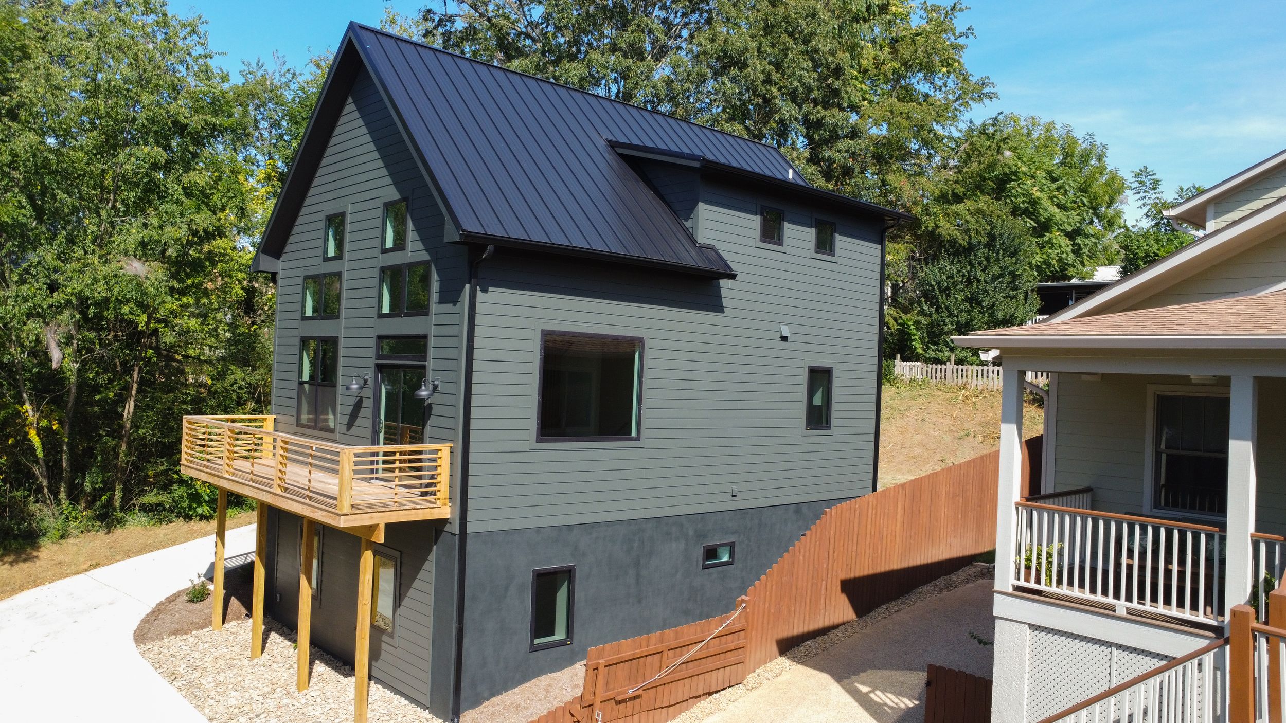 Modern three-story home with gray siding and black metal roof features an elevated wooden deck and walkway.