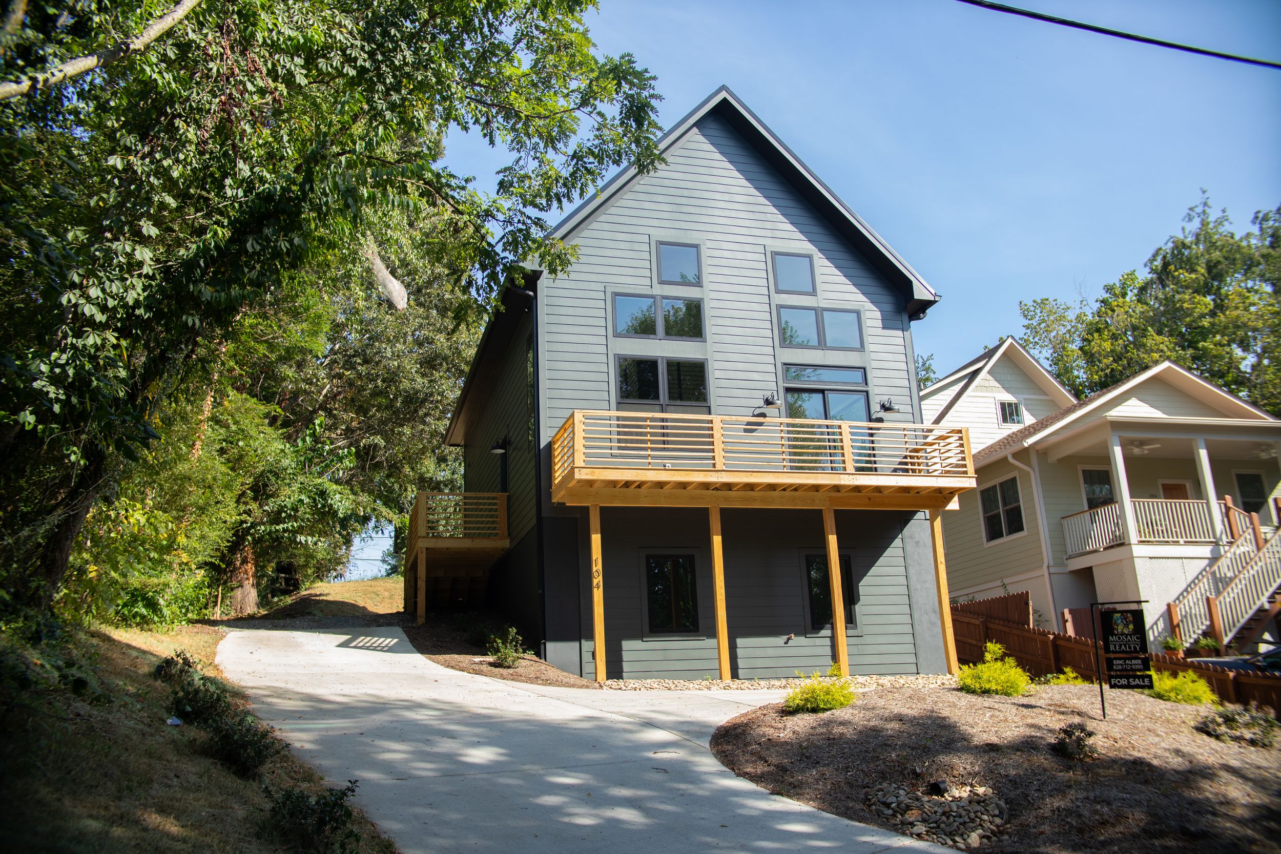 Modern three-story grey house with wood deck balcony built on sloped terrain surrounded by trees.