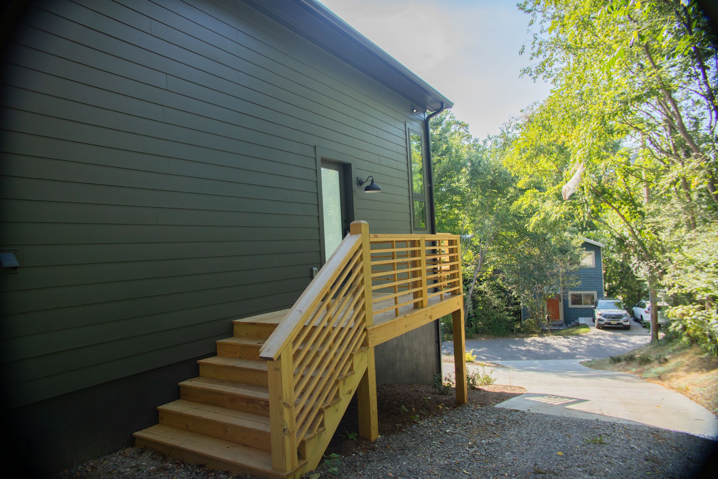 Wooden staircase with modern horizontal railing leads to entrance of dark gray house surrounded by green trees.