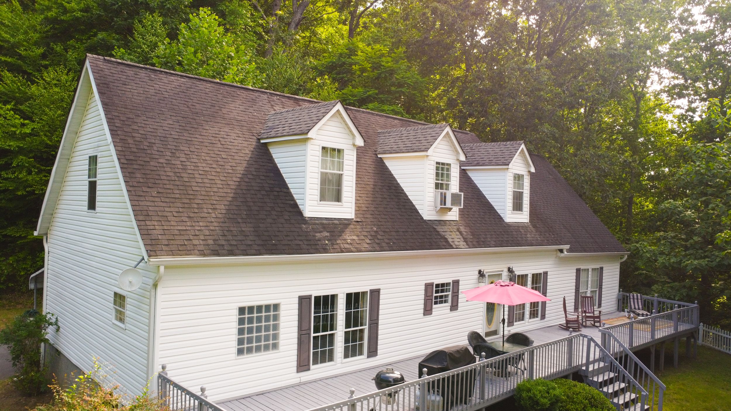 Aerial view of a white Cape Cod style house with three dormers and a large deck surrounded by lush forest.
