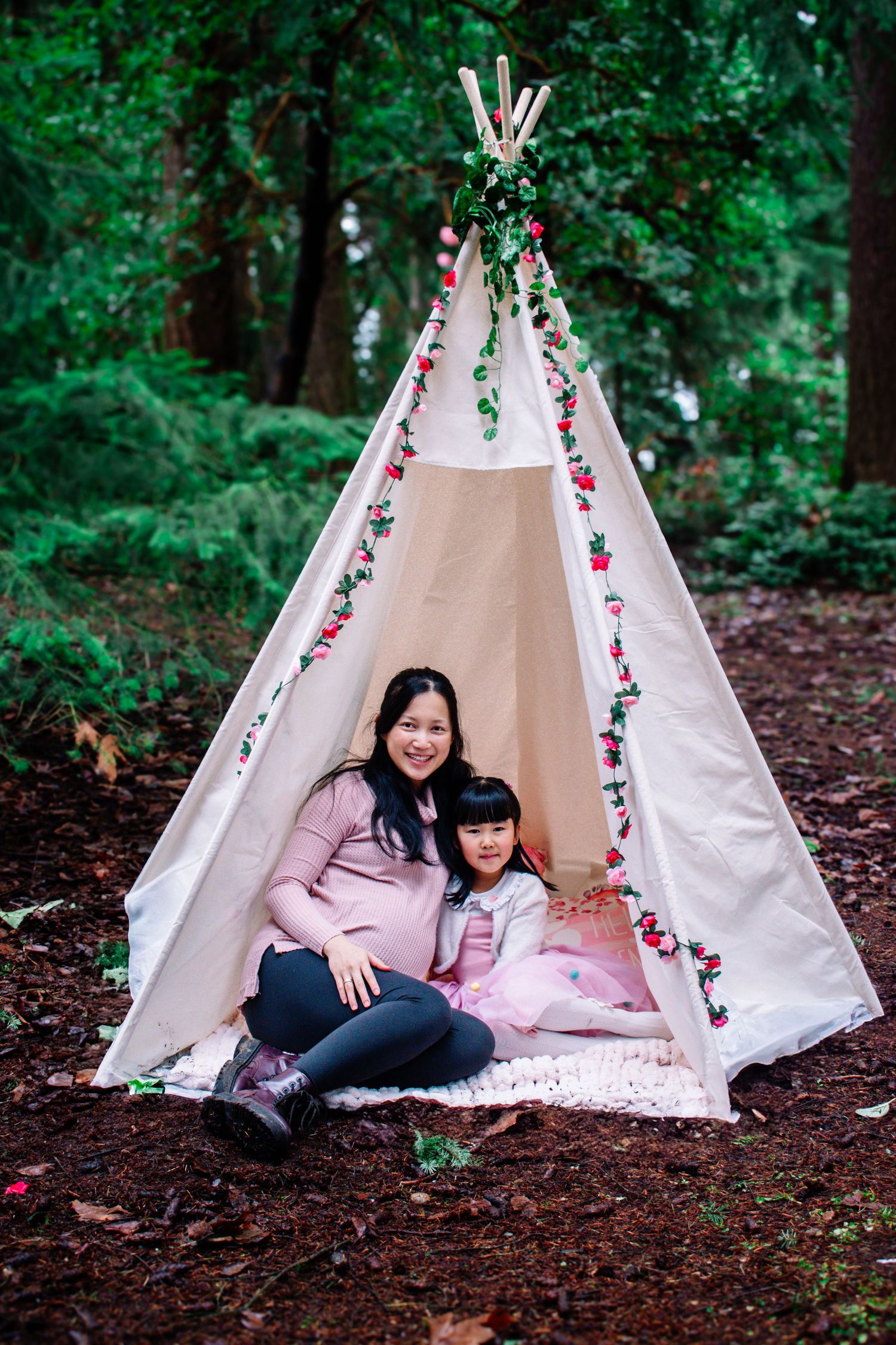A decorated teepee tent with floral garlands sits in a lush green forest setting. Seattle motherhood photography.
