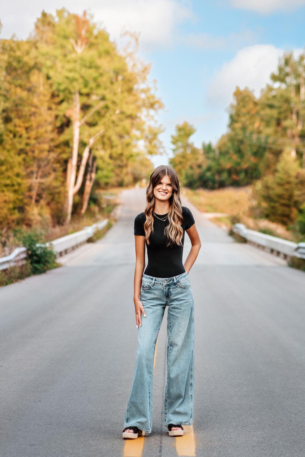 Person wearing black top and light wash wide leg jeans poses casually in middle of empty tree-lined road.