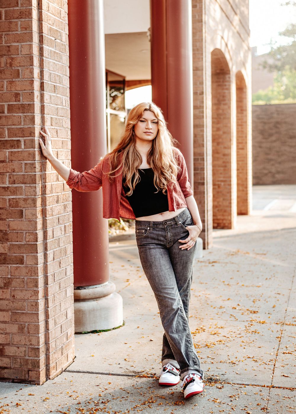 Young woman in a burgundy jacket and jeans leans against a brick column in an outdoor walkway with arches.