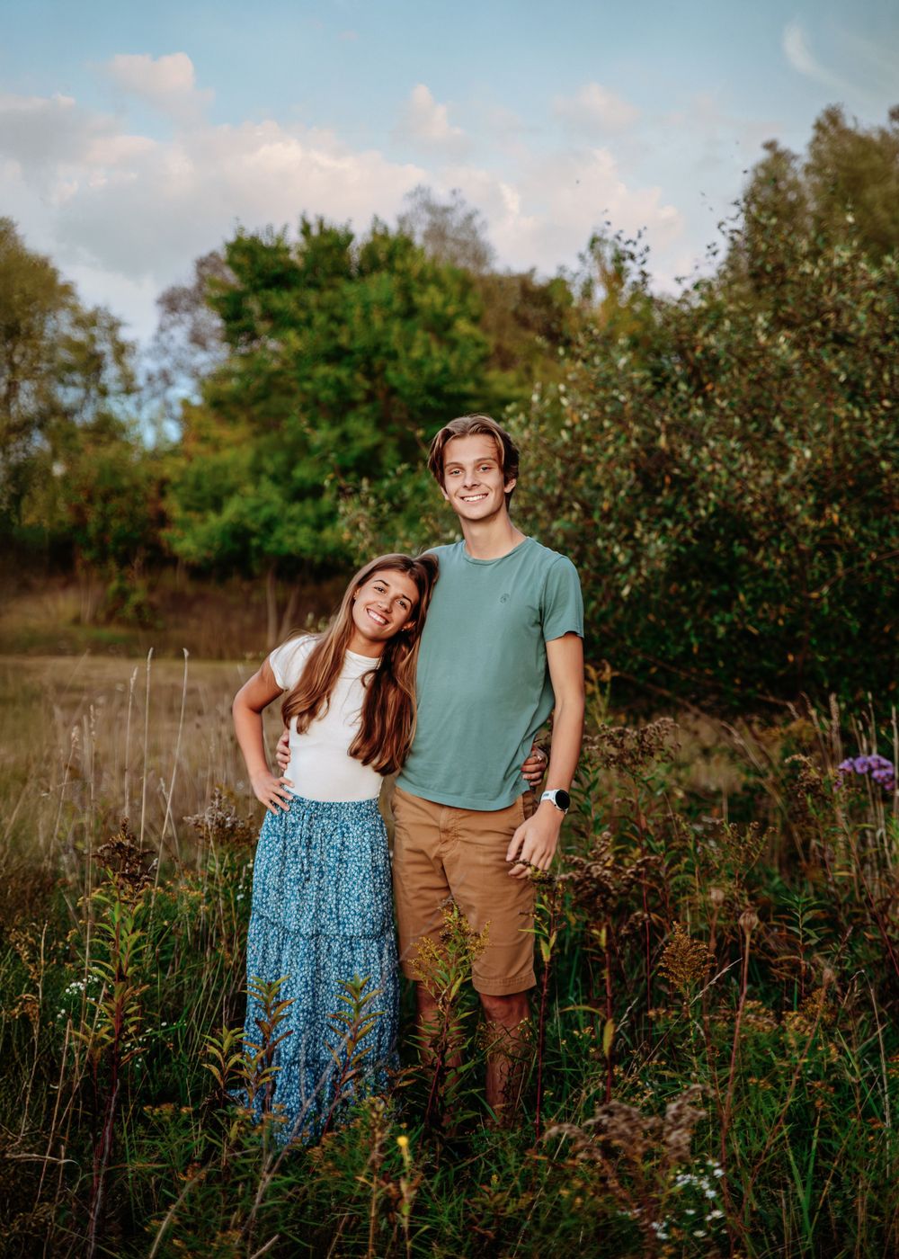 A couple stands together in a wildflower meadow at sunset with trees in the background.