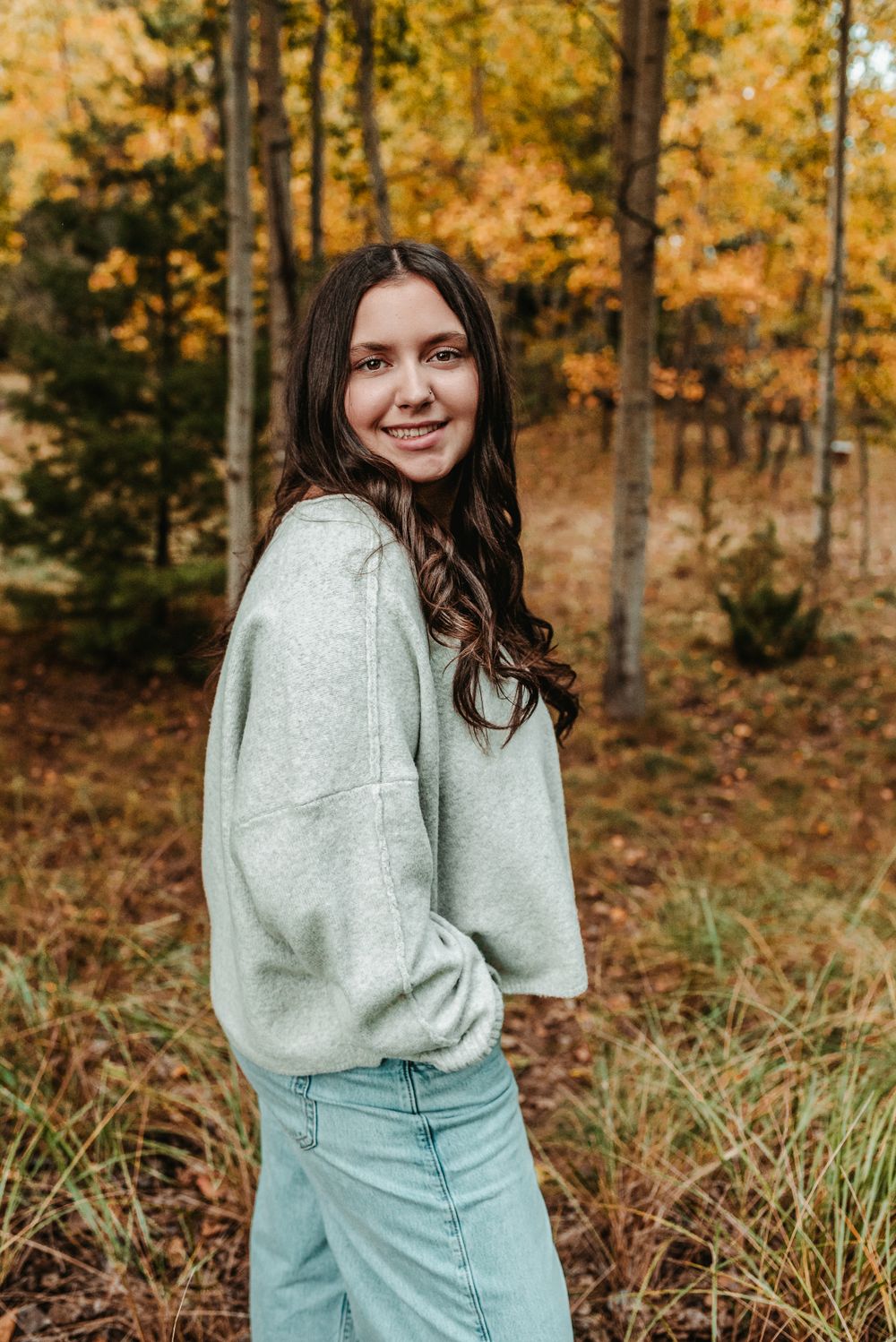 A smiling young person in a light gray sweater and jeans poses in front of golden autumn aspen trees in the forest.